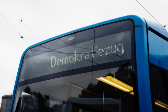 Zu sehen ist der obere Teil einer Straßenbahn von hinten. Auf der digitalen Anzeige ist zu lesen: Demokratiezug.