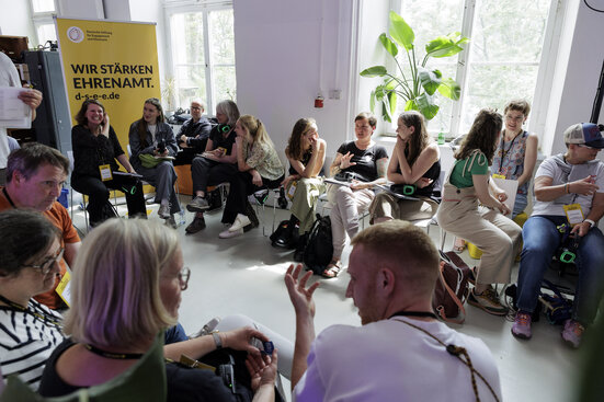 Das Bild zeigt Menschen, die in einem Stuhlkreis angeregt miteinander diskutieren. Im Hintergrund ist großflächig das Logo der Stiftung Engagement und Ehrenamt zu sehen mit dem Slogan: Wir stärken Ehrenamt.