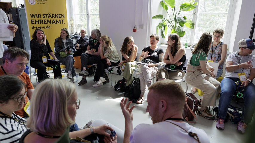 Das Bild zeigt Menschen, die in einem Stuhlkreis angeregt miteinander diskutieren. Im Hintergrund ist großflächig das Logo der Stiftung Engagement und Ehrenamt zu sehen mit dem Slogan: Wir stärken Ehrenamt.