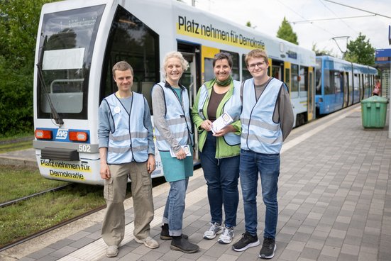 Vier Personen stehen als Diskursbegleiter:innen mit blauen Westen gekennzeichnet, vor dem Demokratiezug, einem Wagen der Straßenbahnlinie 1. 